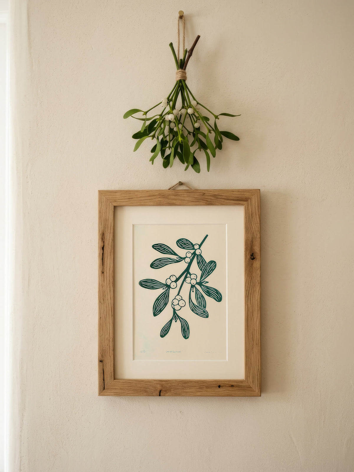 A framed botanical art print of mistletoe hangs on a warm beige wall below a hanging mistletoe sprig. The print features a dark teal-green illustration on an off-white background, enclosed in a light natural wood frame with a white mat.