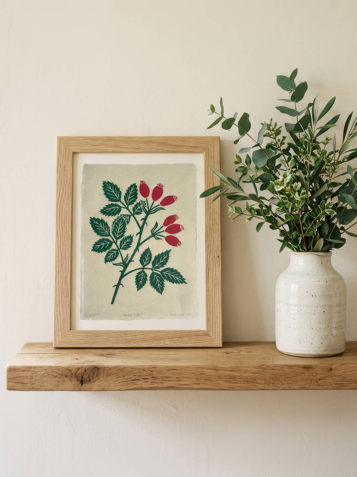 A framed botanical art print featuring green leaves and red berries on a light beige background, styled on a wooden shelf next to a ceramic vase with eucalyptus.
