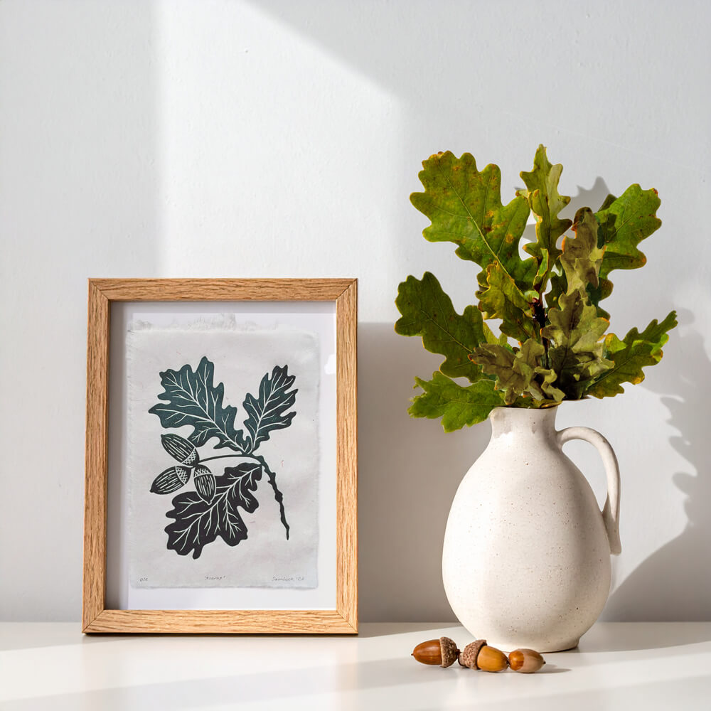Framed botanical artwork of leaves next to a vase with oak leaves and acorns. Sam Luck 6 x 8 inch linocut print wall art, UK.