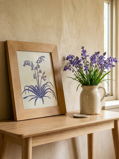 Framed botanical artwork of blue British bluebells on a wooden table with a vase of wild bluebell flowers.