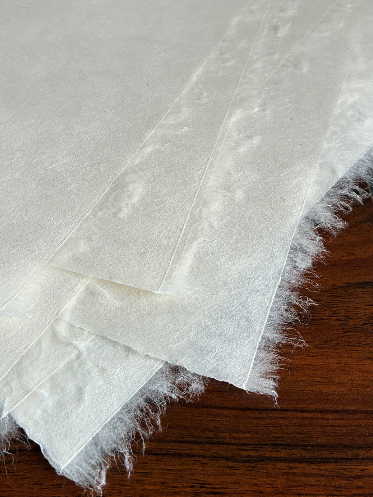 White textured handmade paper with frayed edges layered diagonally on a dark brown wooden surface.