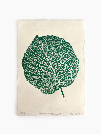 A paper print featuring a green hazel leaf linocut design with white vein details on an off-white textured paper background.