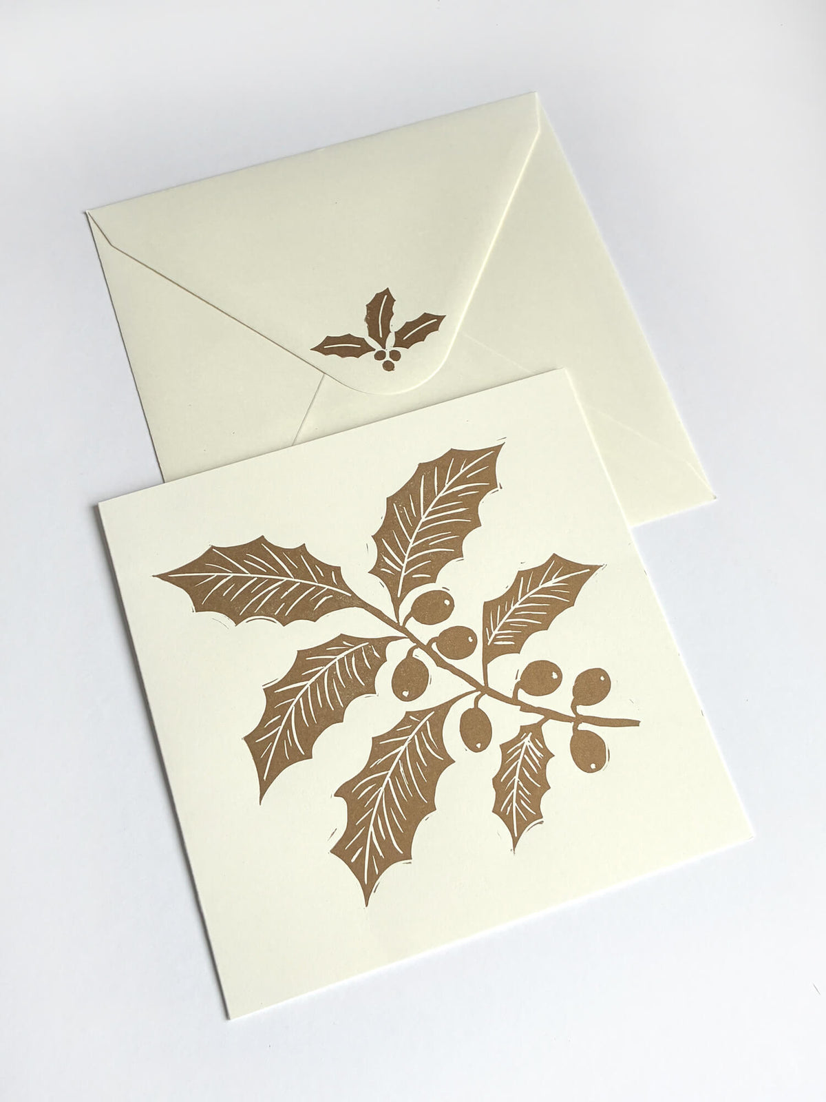 A square cream greeting card with a gold holly branch and berry print, shown with a matching cream envelope on a light grey background.