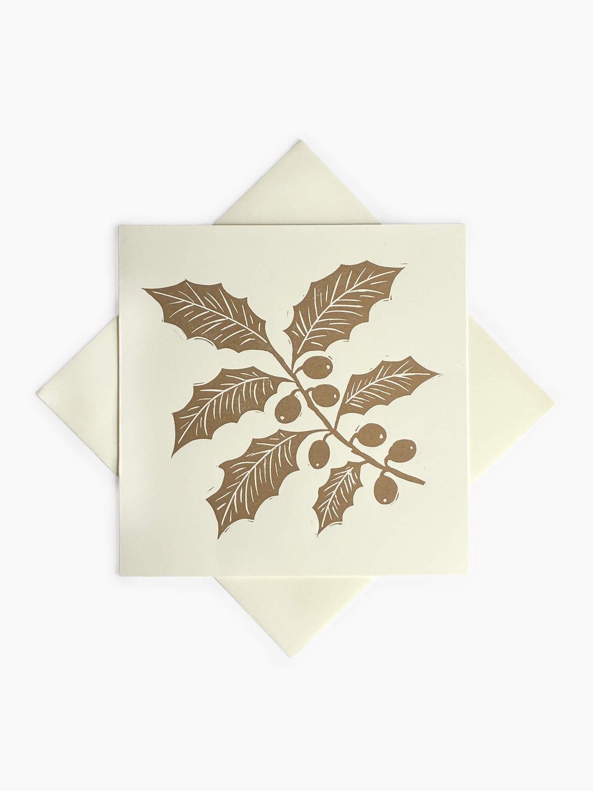 A square cream-colored greeting card featuring a gold holly leaf and berry illustration, layered over a matching cream envelope on a light gray background.