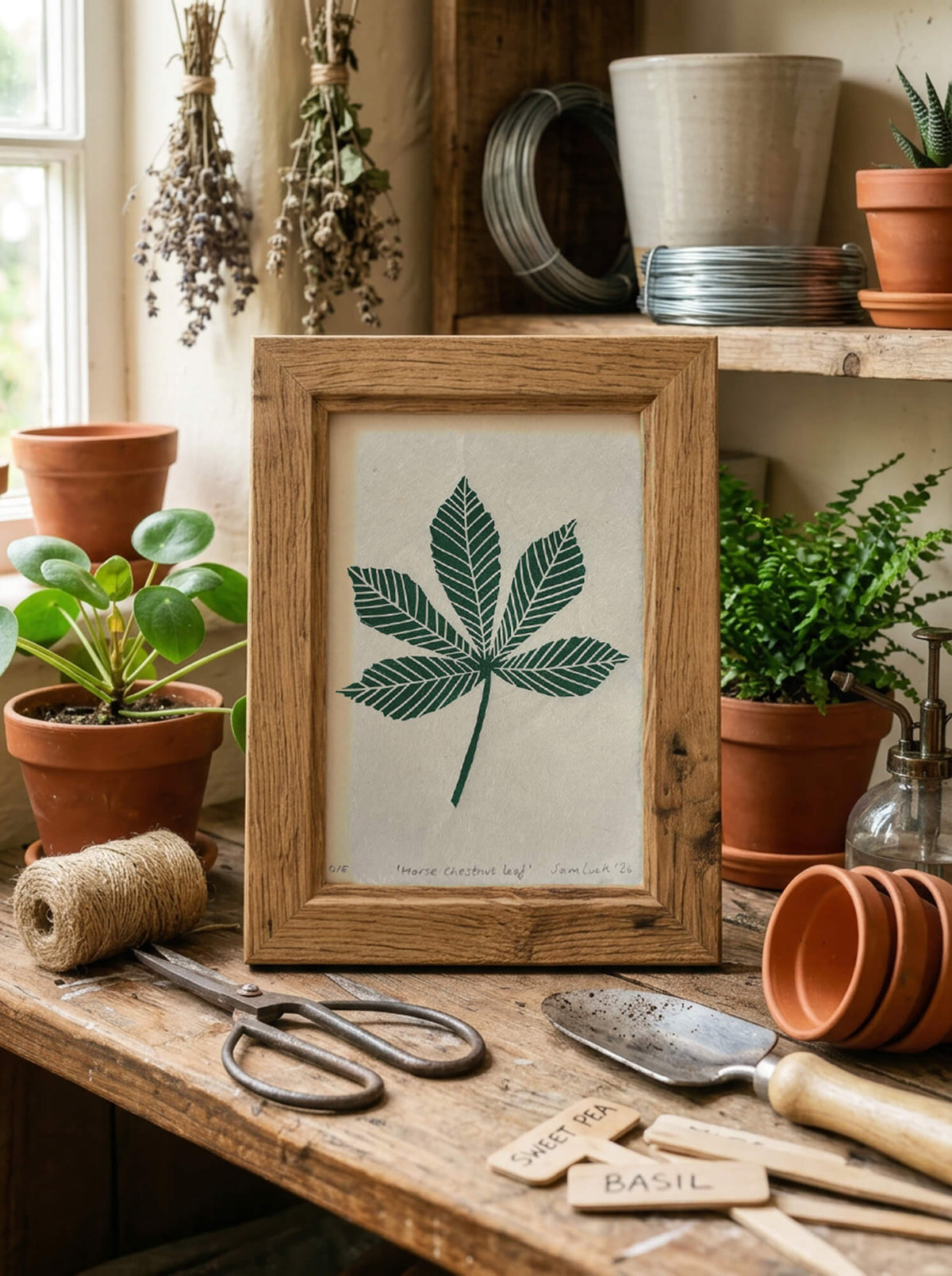 A framed botanical art print featuring a green horse chestnut leaf illustration on an off-white background, styled on a rustic wooden table with gardening tools, potted plants, and dried flowers.