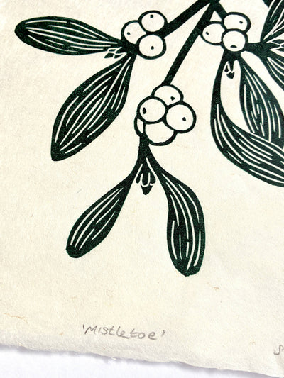 A close-up of a printed botanical design showing mistletoe branches with dark green leaves and white berries on an off-white textured background, with the handwritten word 'Mistletoe' near the bottom.