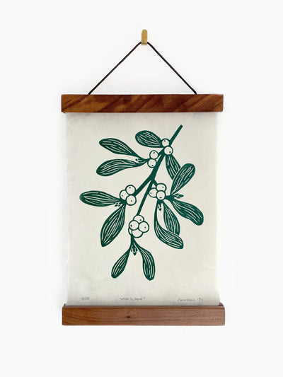A hanging wall art print featuring a green mistletoe botanical illustration on handmade paper hanging from a wooden frame on a white wall.