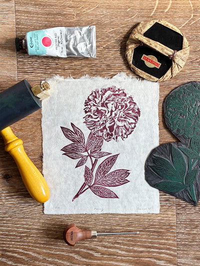 Handmade art print showing a red peony flower block print on off-white handmade paper, surrounded by traditional printing tools including a wooden roller, ink tube, and lino block on a wooden tabletop.