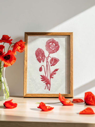 A framed botanical linocut art print featuring red poppy flowers on a textured off-white background, set in a light wood frame. The frame is styled on a light wooden table next to a glass vase of red poppies with scattered petals in the foreground.