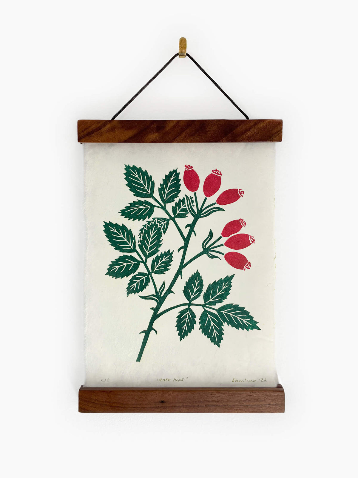 A hanging botanical art print featuring a green rose hip branch with red berries on off-white handmade paper, framed by a dark wood top and bottom bar with a black hanging cord.