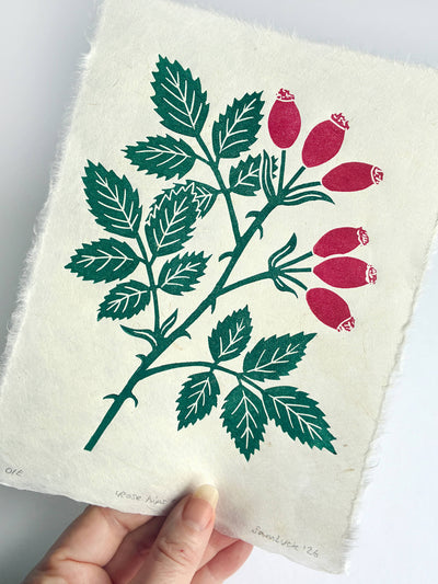 A hand holds a piece of textured off-white paper featuring a botanical print of a rose branch with green leaves and red rose buds, against a plain white background.
