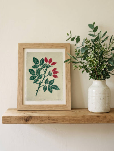 A framed botanical art print featuring green leaves and red berries on a light beige background, styled on a wooden shelf next to a ceramic vase with eucalyptus.