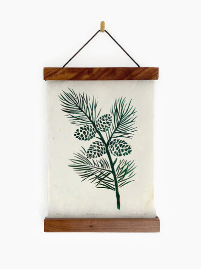 A botanical print featuring a dark green pine branch with pinecones on an off-white handmade paper, suspended from a wooden poster hanging frame on a white wall background.