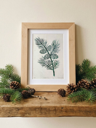 A framed botanical art print of pine branches with green needles and pinecones, displayed on a rustic wooden mantel decorated with pine branches and pinecones against a beige wall.