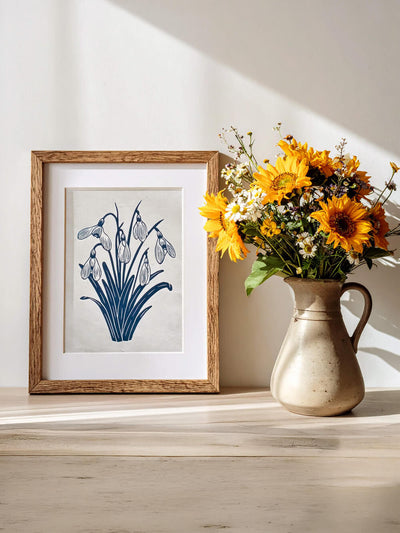 Framed artwork of snowdrops next to a vase with sunflowers and spring flowers on a wooden surface.